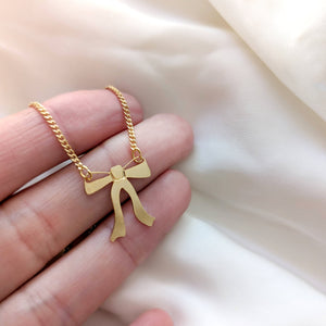 Gold necklace with a ribbon bow pendant hangs from both sides held by fingertips with a white background 