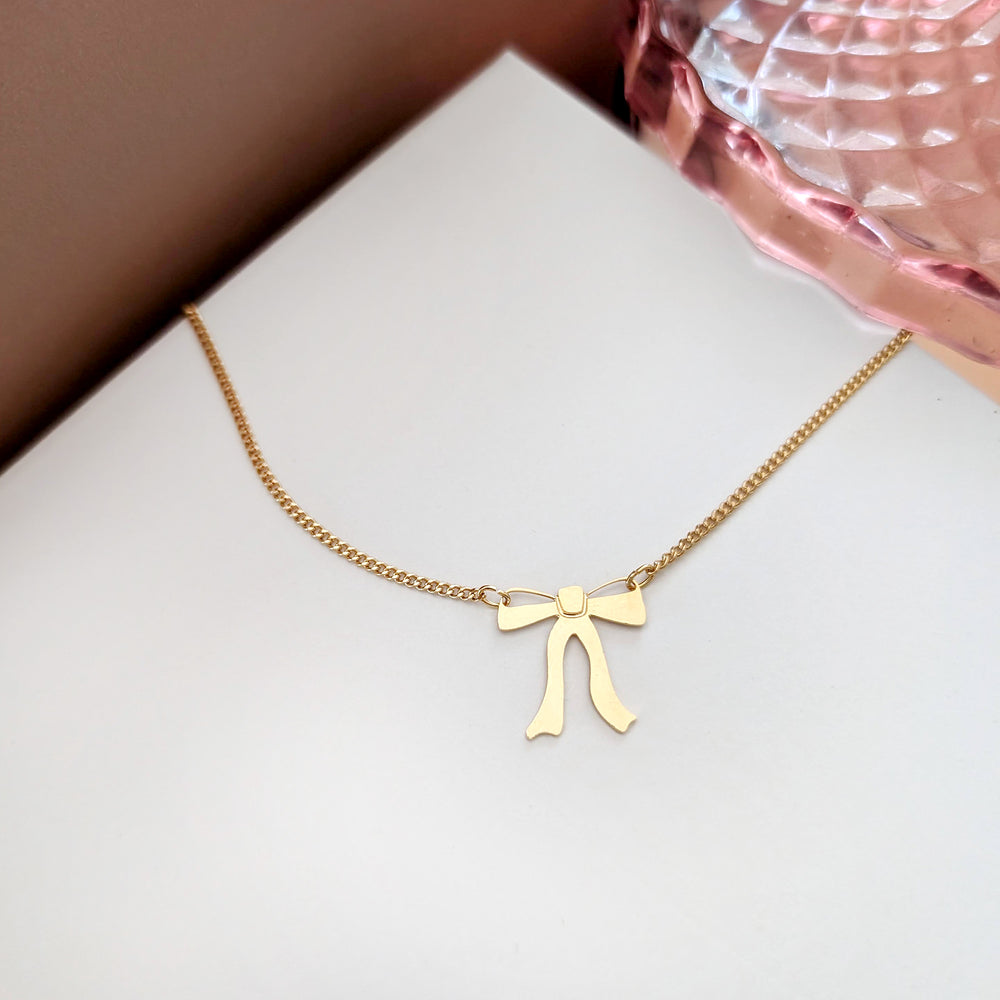 Gold necklace with a ribbon bow pendant hangs from both sides on a white background with a pink glass dish on the side