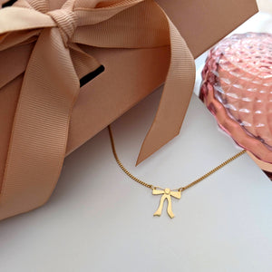 Gold necklace with a ribbon bow pendant hangs from both sides on a white background with a pink gift box on the side