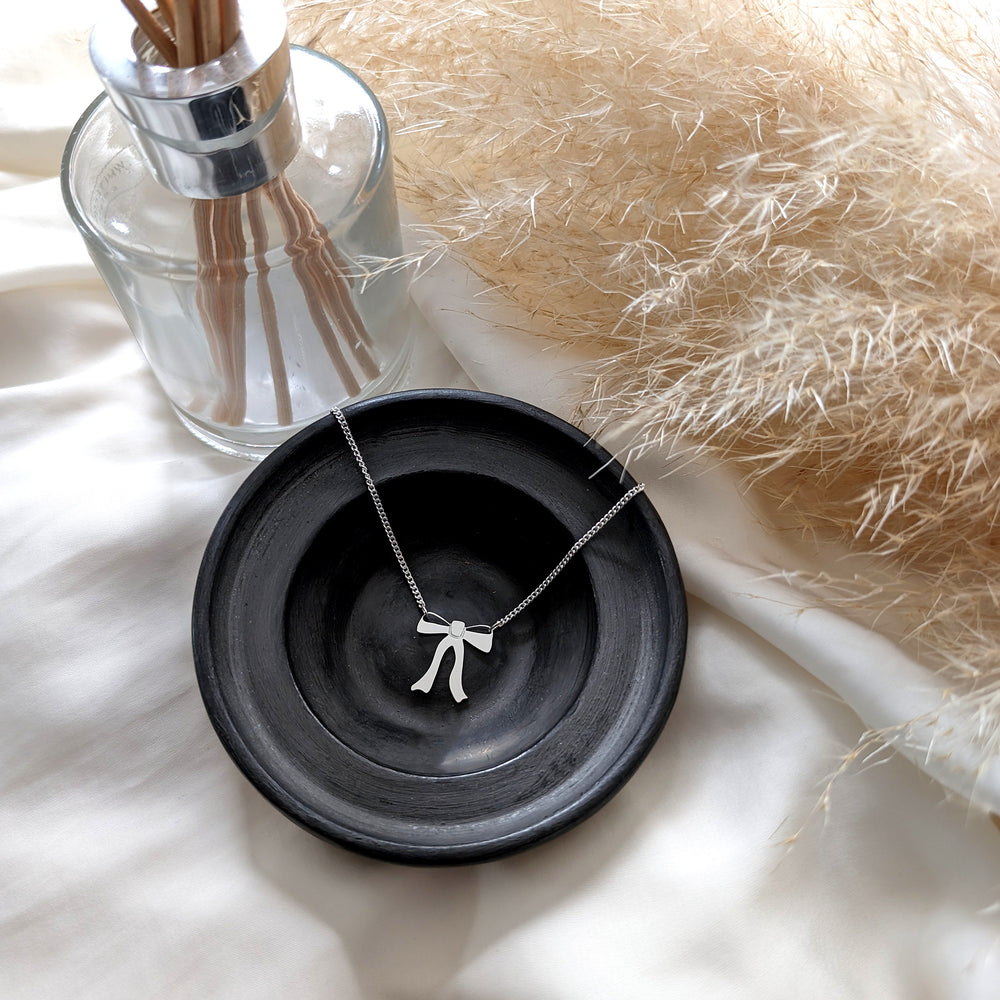 Silver necklace with a ribbon bow pendant hangs from both sides, on a black round little dish with pampas plant on the right side and a diffuser on the left side