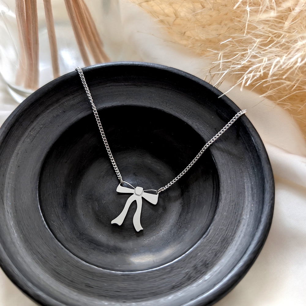  close up on a Silver necklace with a ribbon bow pendant hangs from both sides, on a black round little dish with pampas plant on the right side and a diffuser on the left side